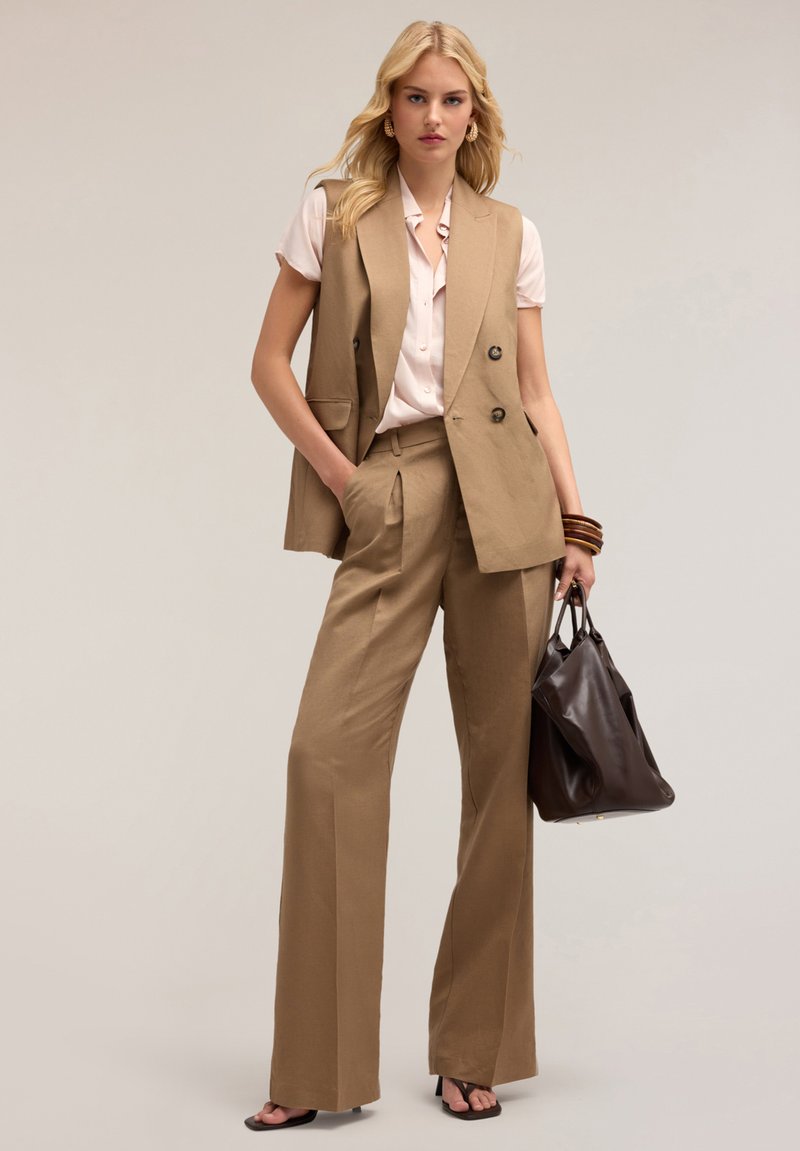 Vaateet, joissa on beige muotolakkovesti ja leveät housut sileästä kankaasta, yhdistettynä vaaleanpunaiseen puseroon. Tummanruskea nahkakäsilaukku ja paksut rannerenkaat.