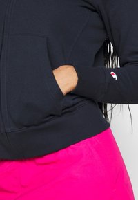 Felpa nera con zip e tasca kangaroo, polsini a coste e piccolo logo sulla manica; indossata con pantaloni corti rosa brillante. Tessuto dalla superficie liscia.