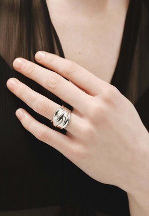 Anillo de plata con un diseño retorcido, que presenta superficies lisas y brillantes, y una banda ancha. Mano descansando sobre una prenda negra texturizada.