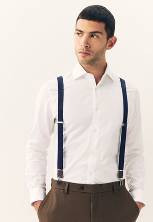 Mann trägt weißes Hemd mit Knöpfen, dunkelblaue Hosenträger und braune Hose, Hände in den Taschen, schaut zur Seite.