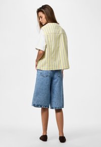 Femme debout, face à l'arrière, portant un gilet jaune à rayures, une chemise blanche à manches courtes, un short en jean à hauteur des genoux et des chaussures slip-on foncées.