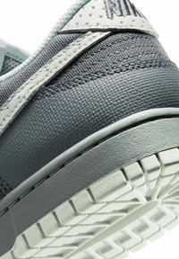 Primo piano del tallone di una sneaker grigia e bianca che mostra tessuto testurizzato, pannello in pelle cucito e suola in gomma con motivo.