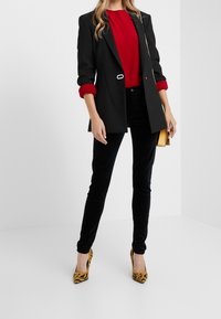 Blazer noir sur mesure avec poignets rouges, accompagné d'un haut rouge et d'un pantalon en velours noir. Des escarpins à motif tigre et une pochette jaune apportent une touche de détail.
