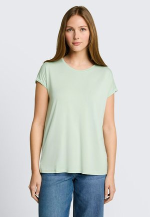 Junge Frau mit glattem hellbraunem Haar trägt ein hellgrünes Kurzarmshirt und blaue Jeans und steht vor einem einfarbigen hellgrauen Hintergrund.