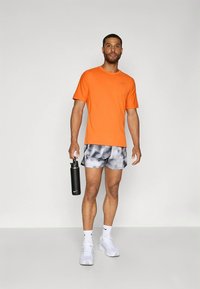 Orange atletisk t-shirt, mönstrade grå shorts, vita sneakers, håller en svart vattenflaska, på en enfärgad bakgrund.