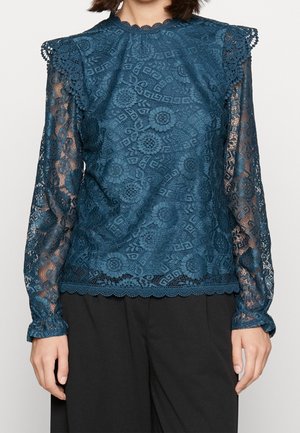 Kobieta ubrana w turkusową koronkową bluzkę w kwiaty z długimi rękawami, marszczonymi ramionami i falbankowym dołem, w połączeniu z czarnymi spodniami.