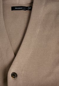 Beige kardigán texturált kötött anyagból, V-nyakkivágással és egy sötét gombbal az elején, címkével a nyakrészben.