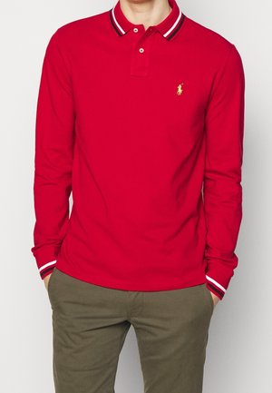 Rode polo met lange mouwen en een klassieke kraag, met een geel logo en gestreepte accenten op de kraag en mouwen.