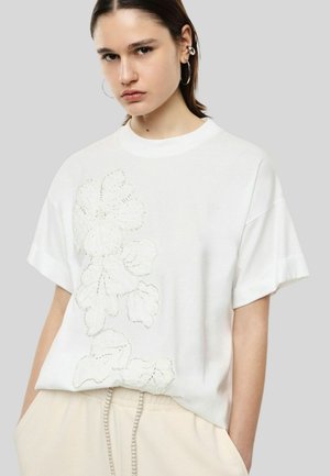 Giovane donna con capelli castani corti che indossa una maglietta bianca oversize con design floreale ricamato e pantaloni beige, in piedi contro uno sfondo grigio chiaro.