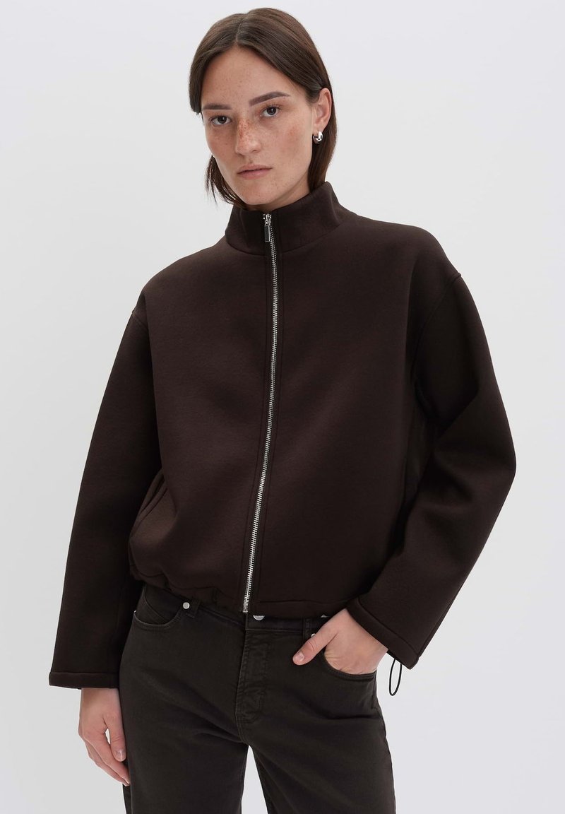 Veste zippée marron en tissu texturé avec un col montant, une coupe décontractée et des poches latérales, associée à un pantalon sombre.