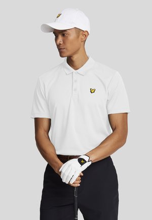 Man som bär vit golfkeps, vit polo med gult fågallogo, vit golfhandske, håller en svart golfklubba, står mot en grå bakgrund.