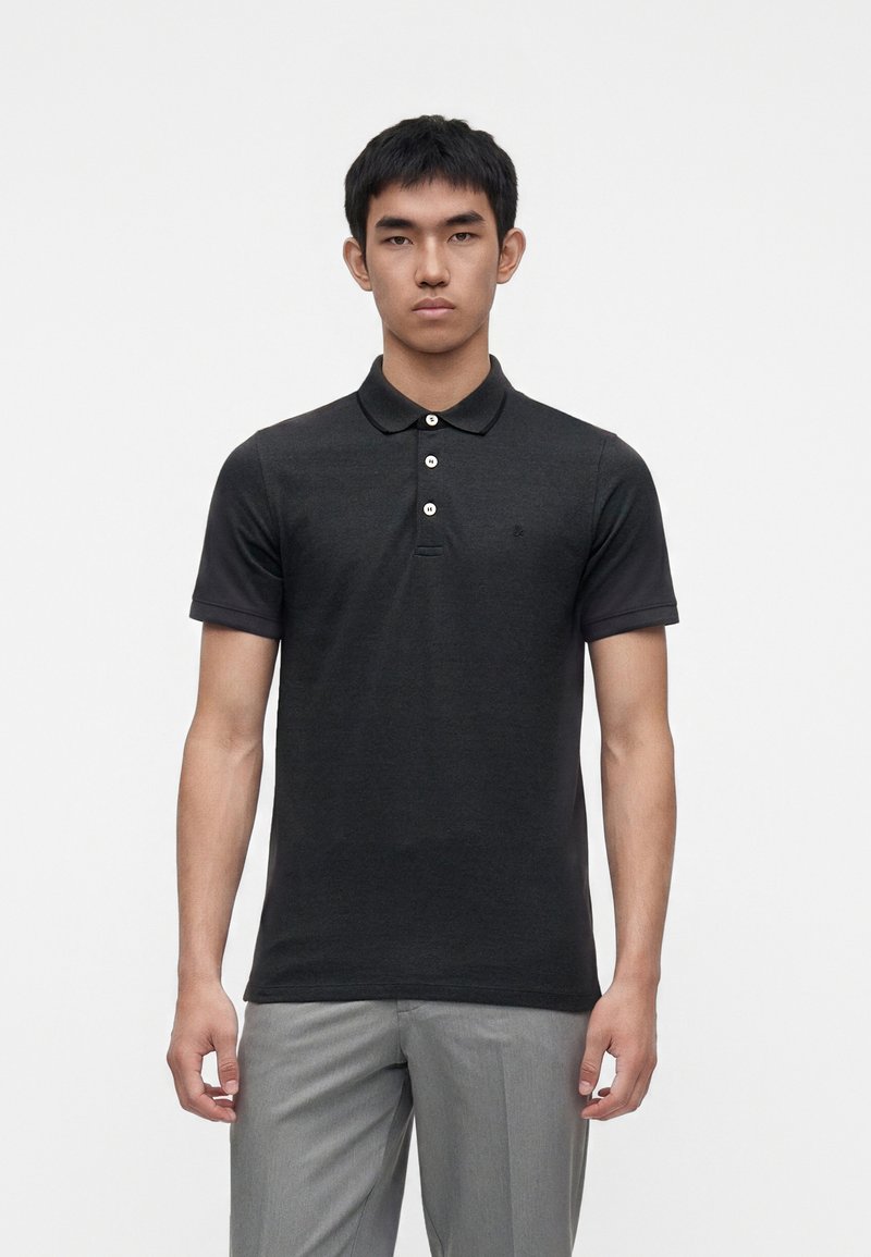 Jeune homme portant un polo noir à manches courtes avec trois boutons et un pantalon gris clair, debout devant un fond blanc uni.