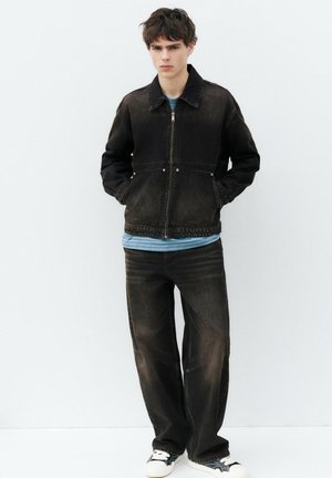 Jeune homme portant une veste en jean noire délavée et un jean, avec une chemise rayée bleue et des baskets noires et blanches, debout contre un mur blanc.