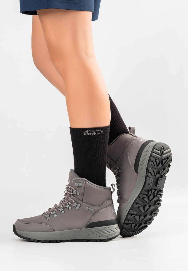 Szare buty trekkingowe z teksturowaną cholewką, wzmocnionymi oczkami i wytrzymałą czarną podeszwą. Noszone z czarnymi skarpetkami w prążki, które sięgają do kostek.