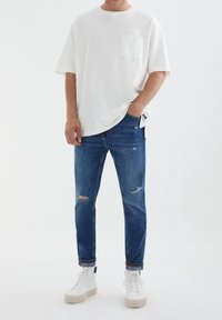 Camiseta blanca de gran tamaño con un bolsillo en el pecho, combinada con jeans azules medios desgarrados. La modelo lleva zapatillas blancas de caña alta. Fondo minimalista.