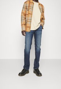 Giacca a scacchi marrone e beige, t-shirt crema, jeans in denim blu e stivali in pelle neri. Bottoni sulla giacca, leggera texture visibile.