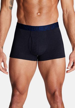 Muskelöser Mann trägt dunkelblaue Boxershorts mit gebrandetem Bund, steht mit entspannten Armen an den Seiten vor einem schlichten Hintergrund.