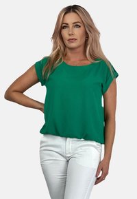 Zielona bluzka z krótkim rękawem, wykonana z gładkiego materiału, z okrągłym dekoltem i luźnym krojem, w zestawie z białymi dopasowanymi dżinsami.