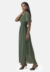 Robe maxi fluide vert olive avec des manches courtes à volants, taille smockée et tissu transparent; accompagnée de sandales à talons hauts noires à lanières.