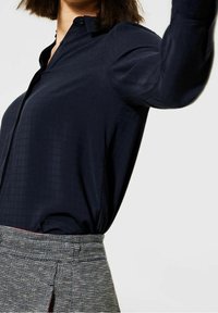 Camicia blu navy realizzata in tessuto morbido, con un motivo a quadretti discreto, colletto abbottonato e maniche lunghe, abbinata a una gonna grigia strutturata.