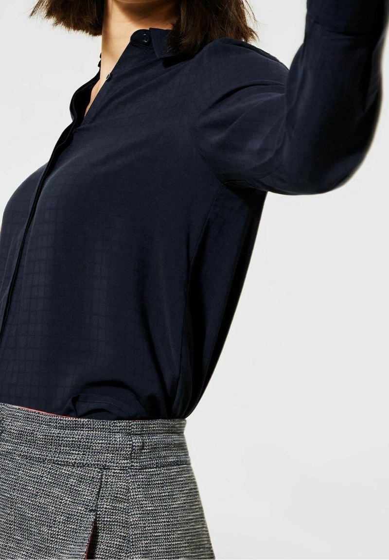 Camicia blu navy realizzata in tessuto morbido, con un motivo a quadretti discreto, colletto abbottonato e maniche lunghe, abbinata a una gonna grigia strutturata.