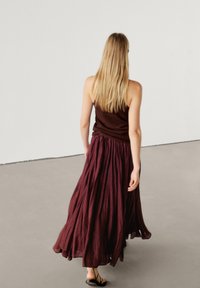 Jupe longue bordeaux avec plis et design fluide, réalisée en tissu léger. Associée à un débardeur moulant marron foncé.