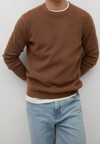 Brązowy sweter o fakturowanej powierzchni z okrągłym dekoltem, w połączeniu z jasnokolorową koszulką pod spodem i niebieskimi dżinsami. Luźny krój, długie rękawy.