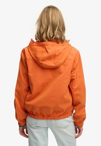 Persona con capelli biondi fino alle spalle che indossa una giacca arancione con cappuccio e jeans azzurri, vista di spalle su sfondo bianco.