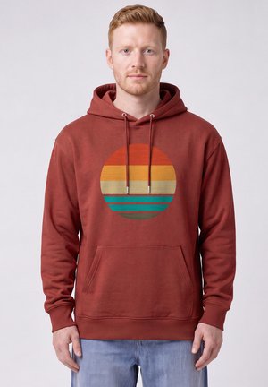 Man draagt een roestkleurige hoodie met een rond retro zonsondergangontwerp in oranje, geel, beige, blauwgroen en bruine strepen, en lichtblauwe spijkerbroek.