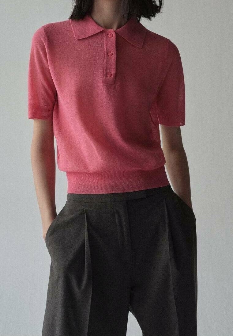 Femme portant un polo rose à manches courtes rentré dans un pantalon noir plissé taille haute, les mains dans les poches.