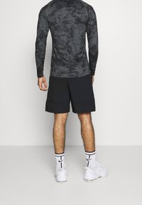 Hombre de pie de espaldas, vestido con pantalones cortos negros, una camiseta de manga larga con un diseño oscuro, calcetines blancos de Nike y zapatillas blancas.