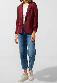 Bordó sako z texturované látky s klopovým límcem a zapínáním na jeden knoflík, doplněno světle modrými džínami a teniskami.