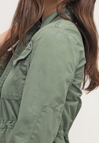 Zielona kurtka z bawełny z dodatkiem innych włókien, z kołnierzykiem, detalami guzików i kieszeniami na piersi. Posiada gromadzony pas, co daje dopasowany kształt.