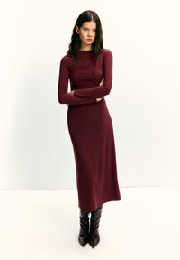 Rochie mulată cu mâneci lungi, de culoare burgundy, confecționată dintr-un material neted, cu decolteu rotund și siluetă până în podea. Purtați împreună cu cizme lucioase până la genunchi.