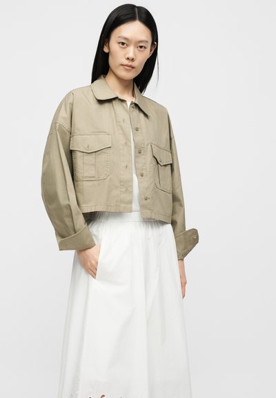 Kvinde med langt sort hår iført cropped beige button-up jakke med store klaplommer over en hvid kjole med sidelommer.