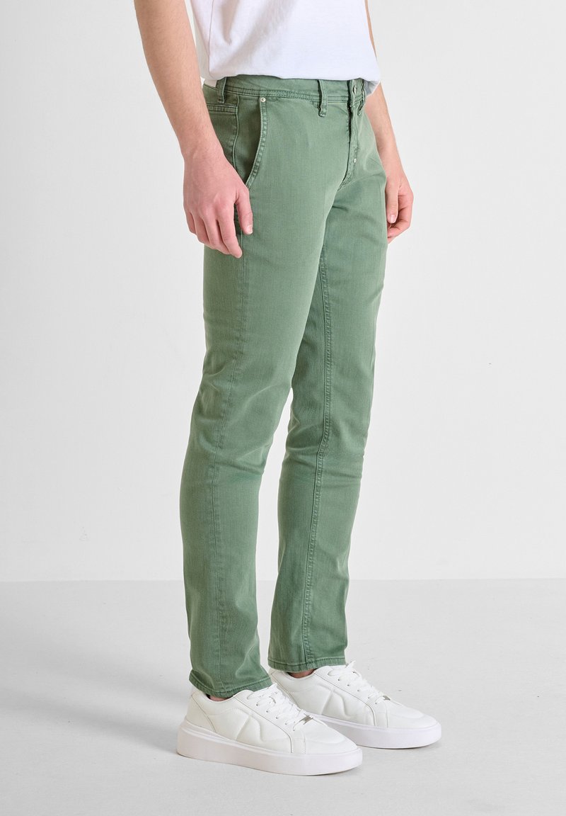 Jeans verdes de algodón de corte slim con bolsillos frontales, costuras en contraste y cremallera, combinados con zapatillas blancas sobre un fondo liso.