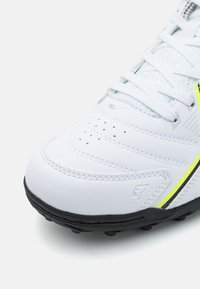 Scarpa sportiva bianca con tomaia sintetica liscia, dettaglio perforato, lacci piatti e suola in gomma nera. Presenta una striscia accentata gialla.