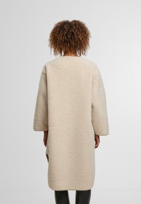 Béžas teksturēts virskrekls ar vaļēju piegriezumu, zemām plecu līnijām un garām piedurknēm, ar sadalītu apakšmalu lielākai kustībai.