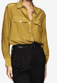 Chemise jaune moutarde à boutons avec deux poches avant, coupe ajustée et texture douce, portée avec un pantalon noir taille haute.