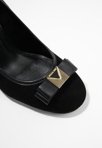 Escarpin en daim noir avec un bout rond, orné d'un nœud en cuir noir et d'un accent triangulaire doré à l'avant. Doublure intérieure lisse.