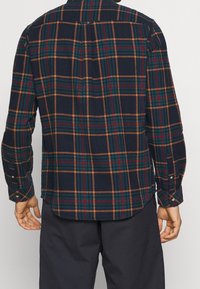 Camisa de franela azul marino con un patrón de cuadros rojos, verdes y amarillos. Manga larga, puños con botones, dobladillo redondeado, detalle en la yugo en la parte posterior.