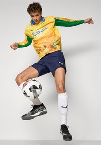 Maglietta a maniche lunghe gialla e verde di Puma con il logo "KidSuper", pantaloni corti blu navy, calze bianche, scarpe da calcio nere; in equilibrio su un pallone da calcio.