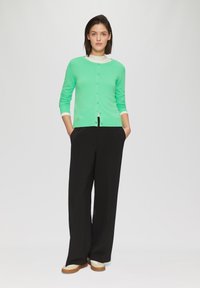 Mintgrüner Cardigan mit rundem Ausschnitt, Knopfleiste und langen Ärmeln; kombiniert mit schwarzen weiten Hosen und Schuhen mit weißer Sohle.