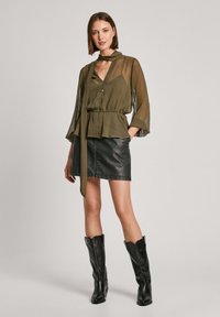 Olive spalvos permatoma blusa su kaklo raiščiu, derinama su juoda odine mini sijonu. Modelis vilki juodus kelių ilgio batus.