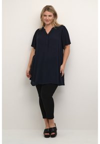 Tunic blu navy a maniche corte con scollatura a V e dettagli di bottoni, abbinata a pantaloni neri aderenti e tacchi aperti neri. Tessuto dalla superficie liscia.