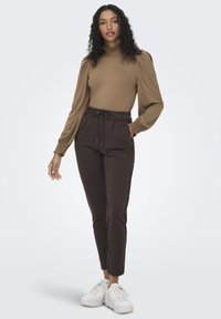 Top marrón de manga larga con cuello y puños con volantes, combinado con pantalones oscuros de corte slim y deportivas blancas. Tejido suave y elástico.