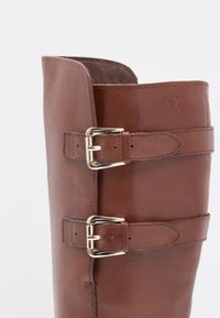 Botte haute en cuir marron avec deux boucles argentées réglables et un petit logo papillon embossé près du haut.