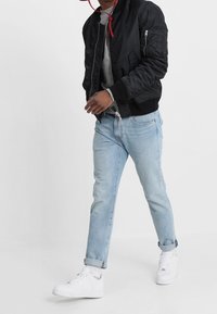 Giacca bomber nera con tasche laterali, felpa grigia sotto, jeans azzurri risvoltati e sneakers bianche, tutto in abbigliamento casual.