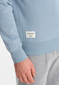 Felpa azzurra chiara con polsini a costine, caratterizzata da un tessuto testurizzato e un'etichetta quadrata del marchio in basso a sinistra con testo stampato.