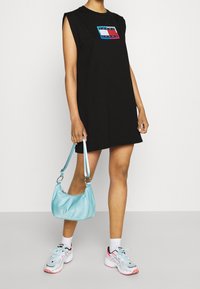 Czarna sukienka bez rękawów z kolorową naszywką logo; zestawiona z miękką, jasnoniebieską torebką na ramię oraz szarymi butami sportowymi z różowymi akcentami.